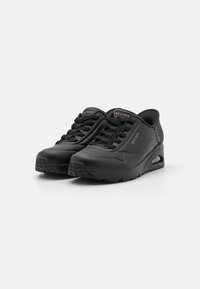 Crne Skechers tenisice s glatkom sintetičkom gornjom stranom, debelim gumene potplatima, okruglim vrhom i izloženom zračnom jastučiću u peti. Crveni vezice s naglascima logotipa.