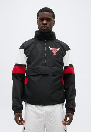 Άνδρας που φοράει αθλητικό μπουφάν σε μαύρο, κόκκινο και άσπρο με το λογότυπο των Chicago Bulls, στέκεται μπροστά σε απλό ανοιχτόχρωμο φόντο.