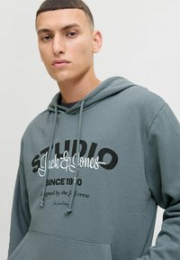 Haut à capuche gris, à enfiler, avec des cordons de serrage, orné d'un grand logo imprimé noir et blanc et de texte sur le devant. Tissu doux, coupe décontractée.