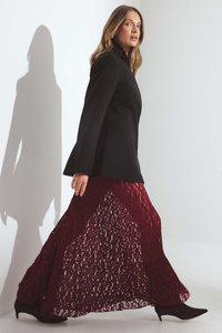 Frau, die in einem schwarzen Blazer, einem bordeauxfarbenen Spitzen-Maxirock und schwarzen Absatzstiefeln vor einem schlichten hellen Hintergrund geht.