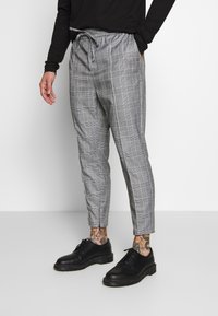 Man wearing gray plaid drawstring pants, black shoes, and a black long-sleeve shirt, standing with one hand in pocket.