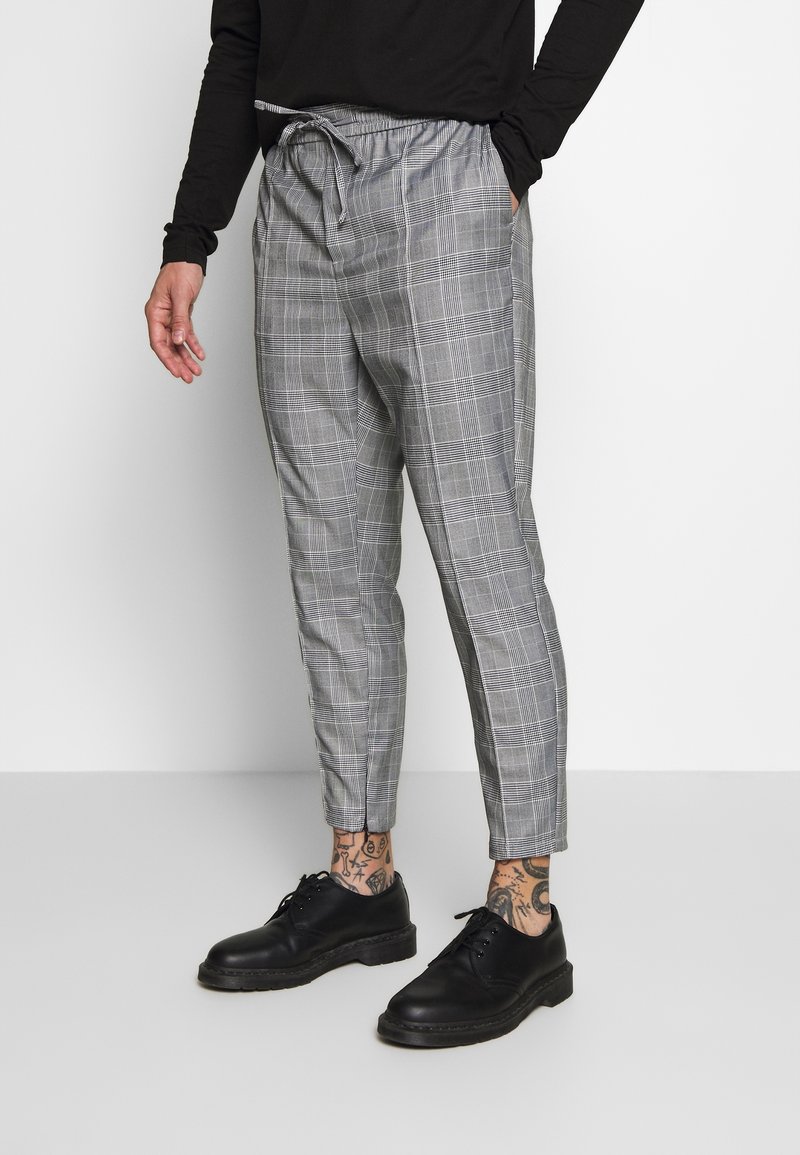 Man wearing gray plaid drawstring pants, black shoes, and a black long-sleeve shirt, standing with one hand in pocket.