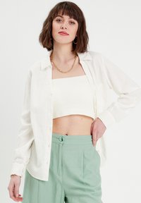 Chemise blanche en lin à boutons sur un crop top crème côtelé, associée à un pantalon taille haute vert clair, présentant un détail de boutons à l'avant.