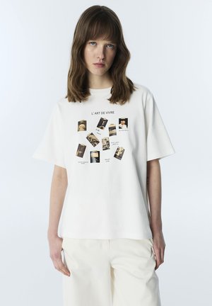 Jeune femme aux cheveux bruns portant un t-shirt blanc avec de petites impressions photo vintage et le texte "L'ART DE VIVRE" sur le devant.