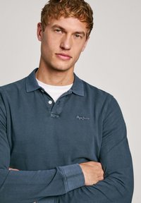 Mörkblå långärmad polo med krage, med en bröstlogotyp och synliga sömmar på ärmsluten och kragen.