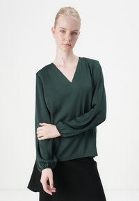 Donkergroen blouse met lange mouwen, een V-hals en geplooide manchetten. Zachte, gladde stof met een relaxte pasvorm en een split aan de zijkant.