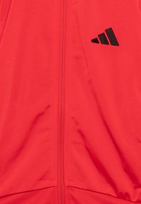 Rote Jacke mit Reißverschluss vorne, glatter Textur und schwarzem Adidas-Logo. Der Stoff hat einen subtilen Glanz und eine leichte Ausführung.