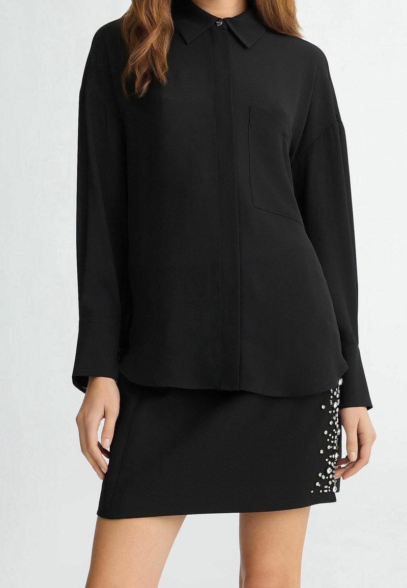 Femme portant une chemise noire à manches longues avec col et une mini-jupe noire ornée de perles sur un côté.