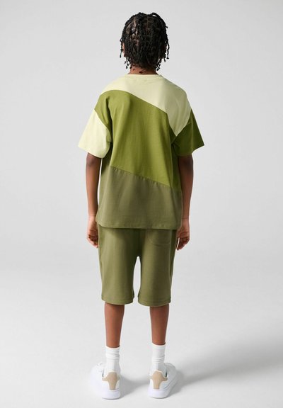 Barn med flettet hår iført grøn T-shirt med blokfarver og matchende shorts, hvide sokker og hvide sneakers, stående mod en ensfarvet baggrund.