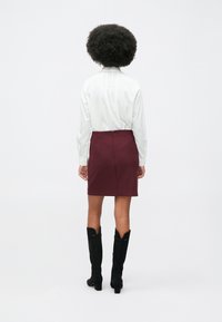 Žena s afro frizurom nosi bijelu majicu dugih rukava, bordo suknju i crne čizme sa petom do koljena, okrenuta leđima na jednostavnoj pozadini.