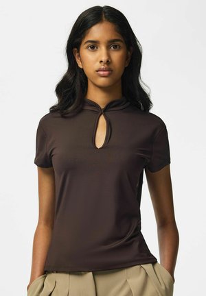 Femme aux longs cheveux foncés portant un haut moulant marron foncé à manches courtes avec une découpe en forme de goutte devant et un pantalon beige, debout devant un fond uni.