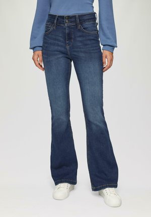 Jean bleu évasé en denim avec une taille haute, doté d'une fermeture à double bouton et de poches avant, associé à des baskets blanches.