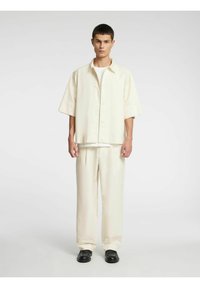 Lys beige kortermet skjorte med myk tekstur, knappestolp foran, og matchende vide bukser. Sorte sko fullfører antrekket.