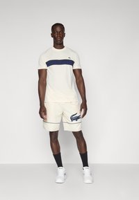 Cream T-shirt with navy horizontal stripe, cream shorts featuring navy crocodile logo, paired with black socks and white sneakers.