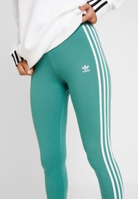 Närbild av gröna Adidas leggings med vit tre-stripes-design och logotyp, som bärs tillsammans med en vit sweatshirt med svarta ränder på ärmarna.