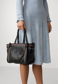 Žena v světle modrých ribbed šatech držící černou koženou kabelku s barevným vyšívaným lemem a dvojitými uchy.