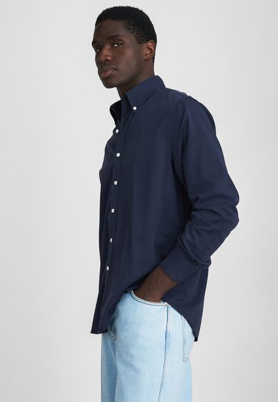 Chemise boutonnée bleu marine à manches longues, avec un col et des boutons blancs. Associée à un denim bleu clair, mettant en avant une coupe décontractée et une texture douce.