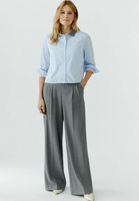 Hellblau gestreiftes Hemd mit Knöpfen und hochgekrempelten Ärmeln; kombiniert mit grauen, hoch taillierten, weit geschnittenen Hosen; glatter Stoff; cremefarbene Schuhe.