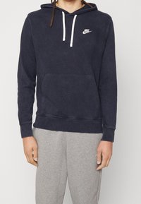 Mörkblå hoodie med dragsko, känguruficka, vitt logo på bröstet, tillverkad av ett mjukt, texturerat tyg. Bärs med grå joggingbyxor.