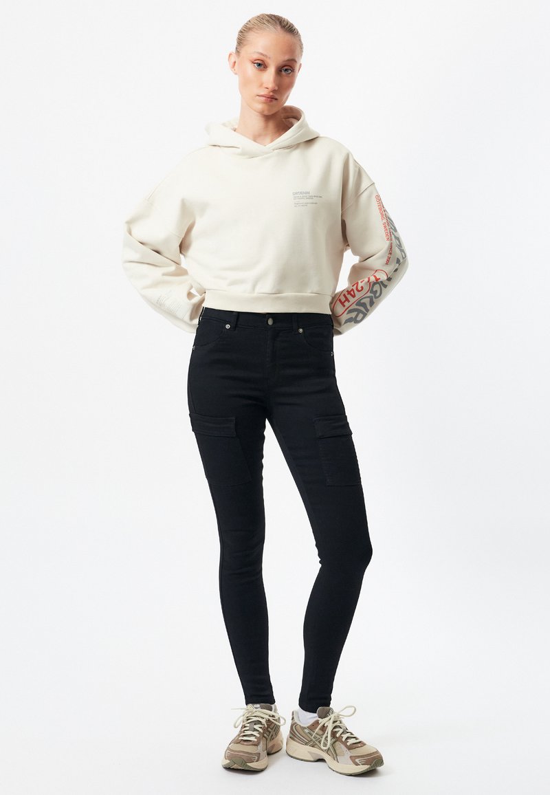 Sudadera corta de color crema con estampados en las mangas; pantalones cargo negros de cintura alta con múltiples bolsillos; zapatillas deportivas de color claro.