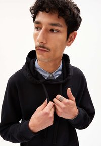 Haut sans manches noir avec une grande capuche et des cordons de serrage, superposé à une chemise à rayures. Tissu lisse, design ajusté et un tatouage subtil visible.