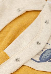 Ropa de algodón con una base de color mostaza y una superposición de beige claro. Presenta botones metálicos y un diseño impreso con acentos azules.