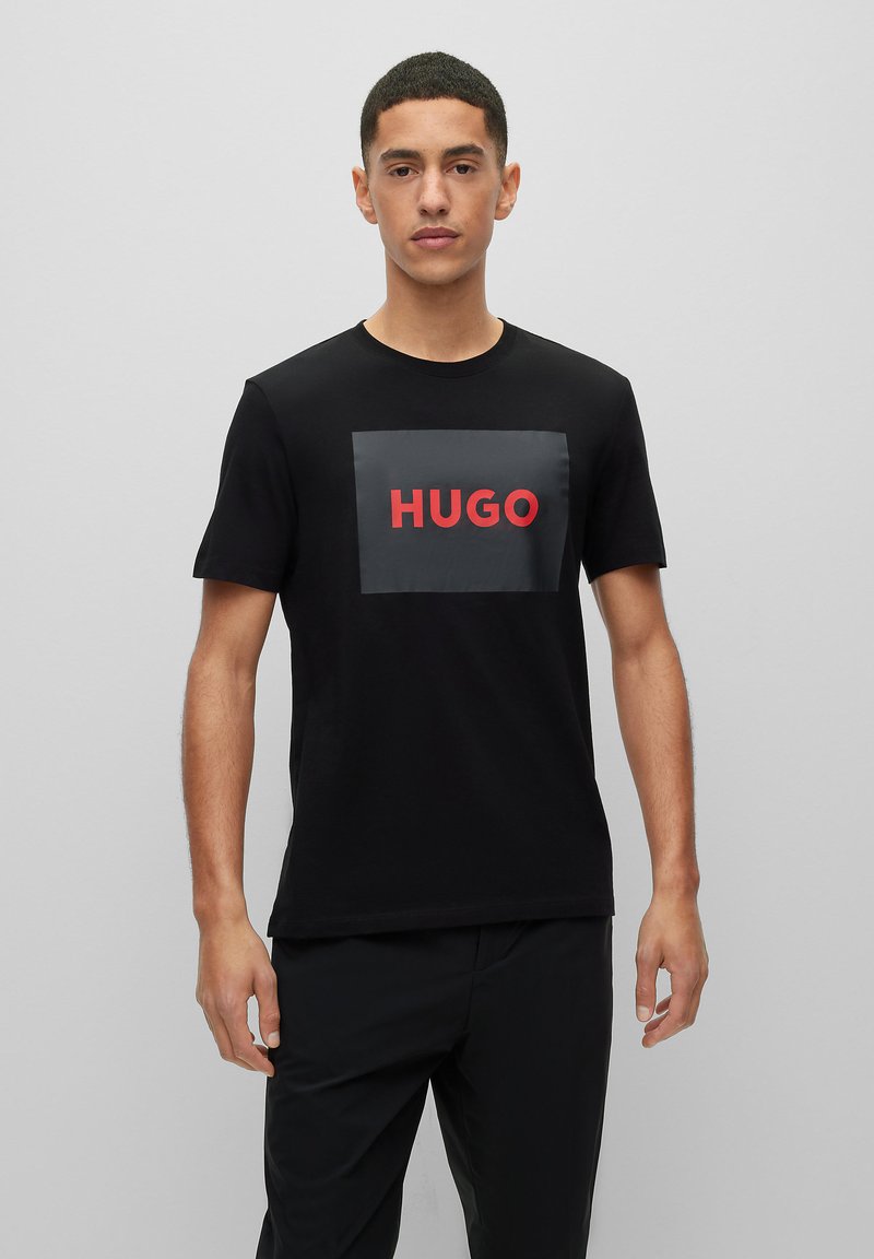 Czarny bawełniany t-shirt z prostokątną szarym naszywką z czerwonym napisem "HUGO" na środku. Krótkie rękawy i okrągły dekolt.
