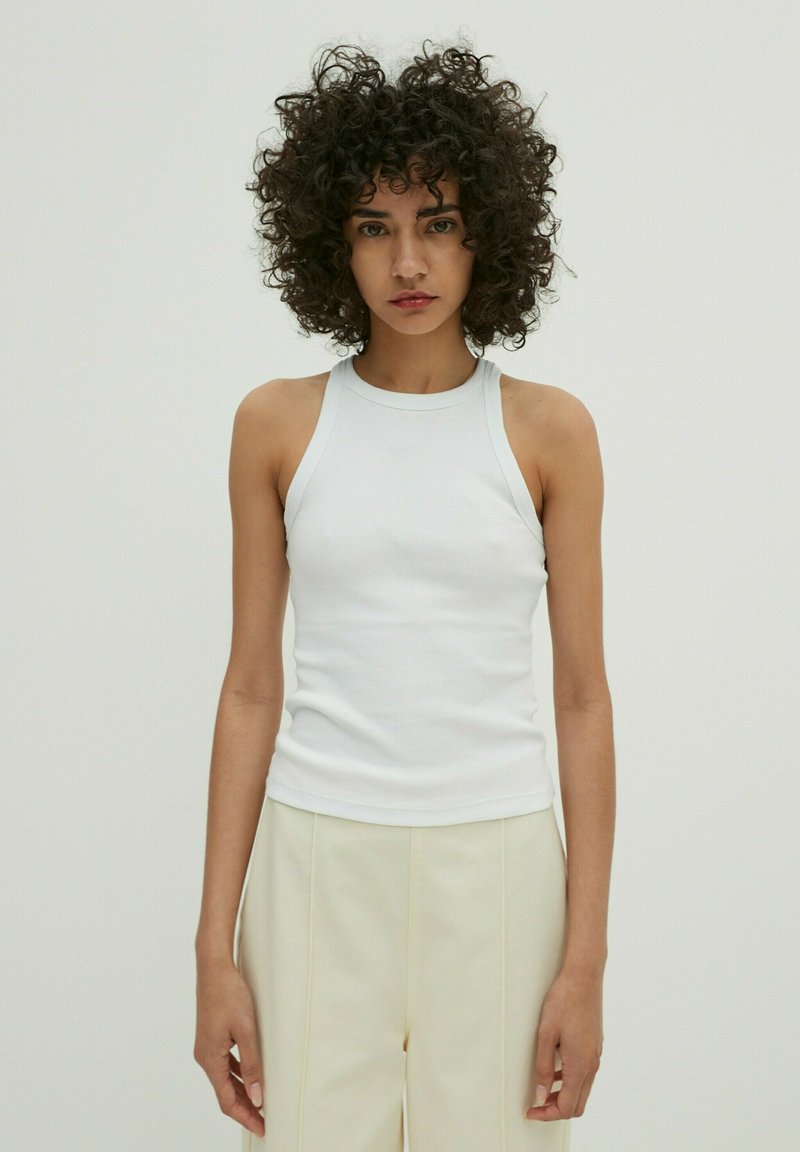 Weiße gerippte Tanktop mit hohem Hals und ärmellosem Design, das eine figurbetonte Form und weiche Textur aufweist, kombiniert mit hellen Hosen.