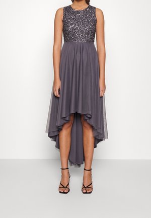 Ärmelloses graues High-Low-Kleid mit glitzerndem Oberteil, fließendem Rock und schwarzen, riemenartigen High-Heel-Sandalen an einer stehenden Person.