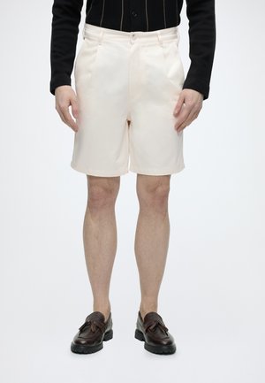 Homme portant un short crème sur mesure, un haut noir à manches longues et des mocassins à pampilles marron foncé, debout devant un fond blanc.
