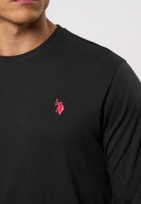 Schwarzes Langarm-T-Shirt aus Baumwolle mit einem kleinen, rosa gestickten Logo auf der linken Brustseite und einem Rundhalsausschnitt.