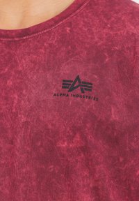 Červené tričko s krátkým rukávem a kulatým výstřihem vyrobené z texturované tkaniny, s černým logem "ALPHA INDUSTRIES" a stylizovanou grafikou na hrudi.