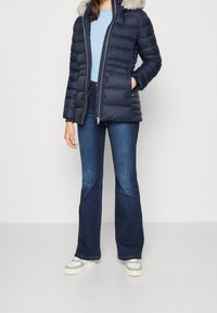 Giacca puffer blu navy con cappuccio bordato in pelliccia, chiusura con zip e polsini a costine, abbinata a jeans neri a zampa d'elefante e scarpe da ginnastica bianche.