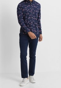 Marineblaue Hemdbluse mit rosa Vogelmuster, Baumwollstoff, langen Ärmeln und Kragen, kombiniert mit marineblauer Hose und weißen Turnschuhen.