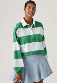 Γυναίκα που φοράει μακρυμάνικο πόλο με πράσινες και άσπρες ρίγες και ανοιχτόχρωμη πλισέ τζιν φούστα, στέκεται μπροστά σε απλό φόντο.