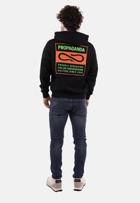 Felpa nera con cappuccio caratterizzata da una grande grafica colorata sul retro con il testo "PROPAGANDA" e un logo, abbinata a jeans blu e sneaker chiare.