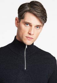 Schwarzer Strickpullover mit hohem Kragen und silbernem Reißverschluss am Hals. Glatte Textur, taillierte Passform und minimalistisches Erscheinungsbild.