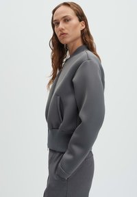 Giacca bomber grigia in tessuto liscio e stretch con orlo a coste, tasche laterali e vestibilità comoda. Design minimalista, senza chiusure visibili.