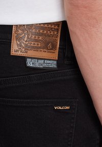 Un jean en denim noir présente une étiquette en cuir marron texturé avec un design graphique et un logo. Une étiquette mentionne les avantages en matière d'économie d'eau et les indicateurs.