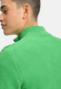 Grön stickad tröja med hög krage, med ett strukturerat mönster och ribbat avslut runt halsen och ärmarna.