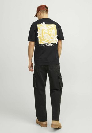 Camiseta de algodón negra con un gráfico floral amarillo y blanco en la espalda, combinada con pantalones cargo negros y una gorra marrón.