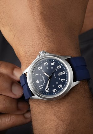 Blue silicone strap watch with a stainless steel case, round face, white Arabic numerals, a date window, and silver detailing on the crown.
