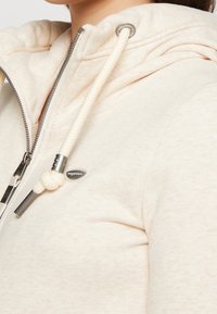 Sudadera con cremallera de color beige claro, hecha de tejido suave. Cuenta con una capucha ajustable, cremallera metálica y una etiqueta de logo en el área del pecho.