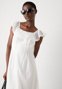 Rochie din bumbac alb cu broderie, cu un decolteu volanat, având motive decupate. Modelul poartă ochelari de soare pătrați negri și cercei rotunzi din argint.