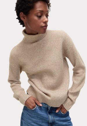 Beige ribbed turtleneck sweater with a relaxed fit and long sleeves, featuring a textured knit and ribbed cuffs and hem. Worn over denim.