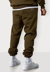 Olivenfarbene Jogginghosen mit schmalem Schnitt, elastischen Bündchen und gestickten Akzenten. Kombiniert mit weißen Turnschuhen und sichtbaren weißen Socken.