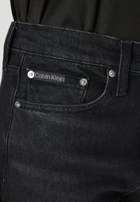 Vaqueros de mezclilla negros con una textura suave, que presentan un parche con el logo de Calvin Klein, herrajes plateados y un diseño clásico de cinco bolsillos.