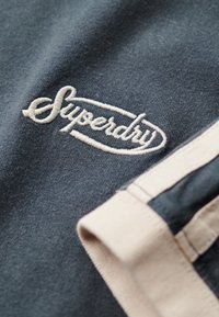 Sudadera azul marino con el logotipo bordado de "Superdry" en crema. Presenta una textura suave y detalles de rayas en beige claro en la manga.