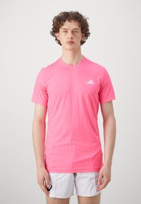 Pink athletic shirt with a textured pattern, short sleeves, crew neck, and a white logo on the left chest. Worn with white shorts.