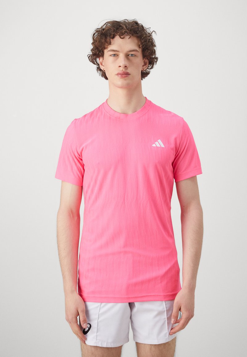 Pink athletic shirt with a textured pattern, short sleeves, crew neck, and a white logo on the left chest. Worn with white shorts.